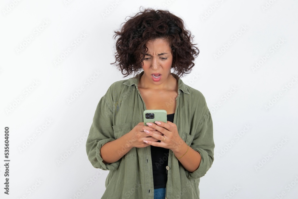 Focused young beautiful woman with curly short hair wearing green overshirt over white wall use smartphone reading social media news, or important e-mail Focused young beautiful woman with curly short hair wearing green overshirt over white wall use smartphone reading social media news, or important e-mail