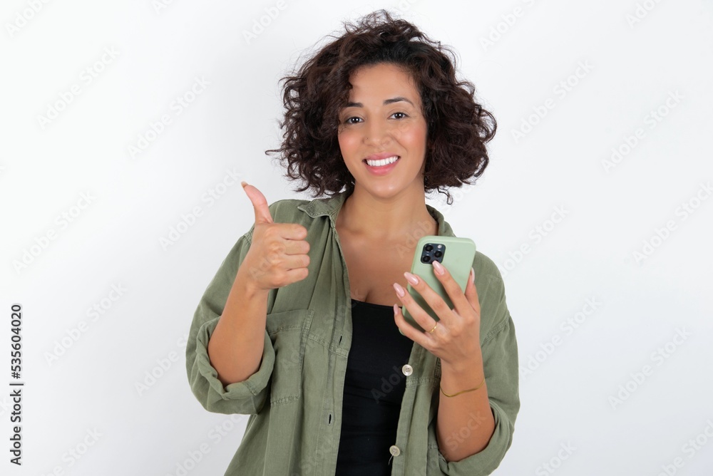 Portrait of young beautiful woman with curly short hair wearing green overshirt over white using and texting with smartphone happy with big smile doing ok sign, thumb up with fingers, excellent sign Portrait of young beautiful woman with curly short hair wearing green overshirt over white using and texting with smartphone happy with big smile doing ok sign, thumb up with fingers, excellent sign