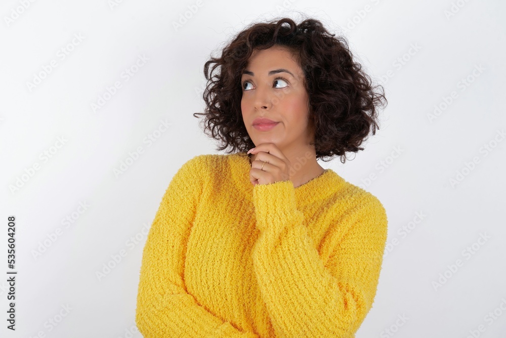 Thoughtful young beautiful woman with curly short hair wearing yellow sweater over white background holds chin and looks away pensively makes up great plan Thoughtful young beautiful woman with curly short hair wearing yellow sweater over white background holds chin and looks away pensively makes up great plan