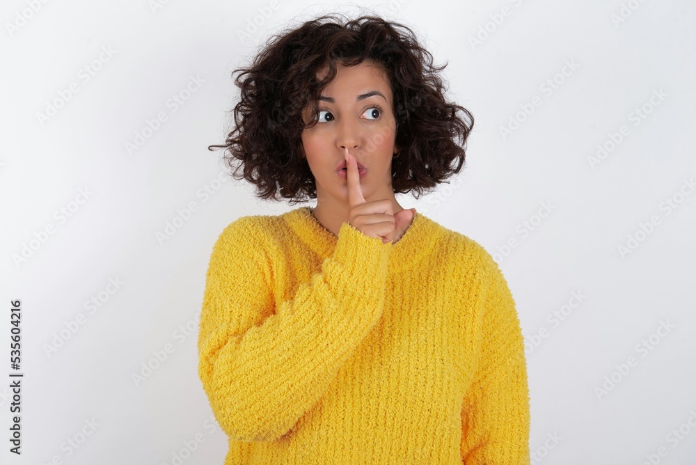 young beautiful woman with curly short hair wearing yellow sweater over white background silence gesture keeps index finger to lips makes hush sign. Asks not to share secret young beautiful woman with curly short hair wearing yellow sweater over white background silence gesture keeps index finger to lips makes hush sign. Asks not to share secret