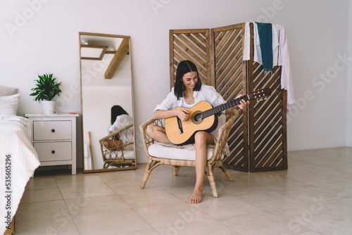 Brunette attractive female smiling and learning to play guitar while sitting in cozy home interior. Young happy woman enjoying her hobby with musical instrument performance relaxing on wicker chair