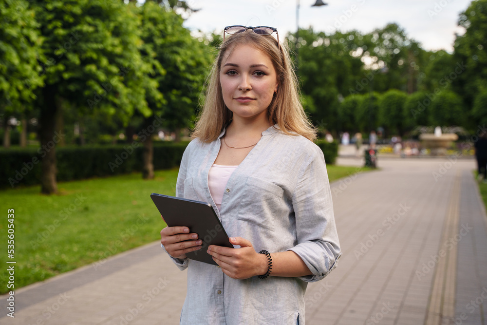 Fototapeta premium Portrait of young female student holding tablet in summer public park in sunny day.