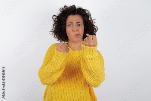 Displeased annoyed young beautiful woman with curly short hair wearing yellow clenches fists, gestures pissed, ready to revenge, looks with aggression at camera stands full of hate, being pressured photo