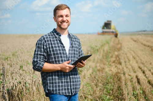 Happy farmer proudly standing in a field. Combine harvester driver going to crop rich wheat harvest. Agronomist wearing flannel shirt, looking at camera on a farmland