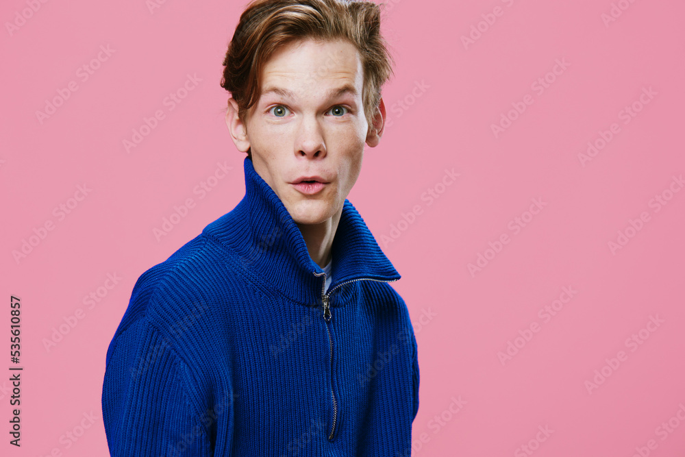 funny playful red-haired young man stands in a blue sweater with a zipper and makes a funny face while looking at the camera. Horizontal studio portrait on pink background with empty space funny playful red-haired young man stands in a blue sweater with a zipper and makes a funny face while looking at the camera. Horizontal studio portrait on pink background with empty space