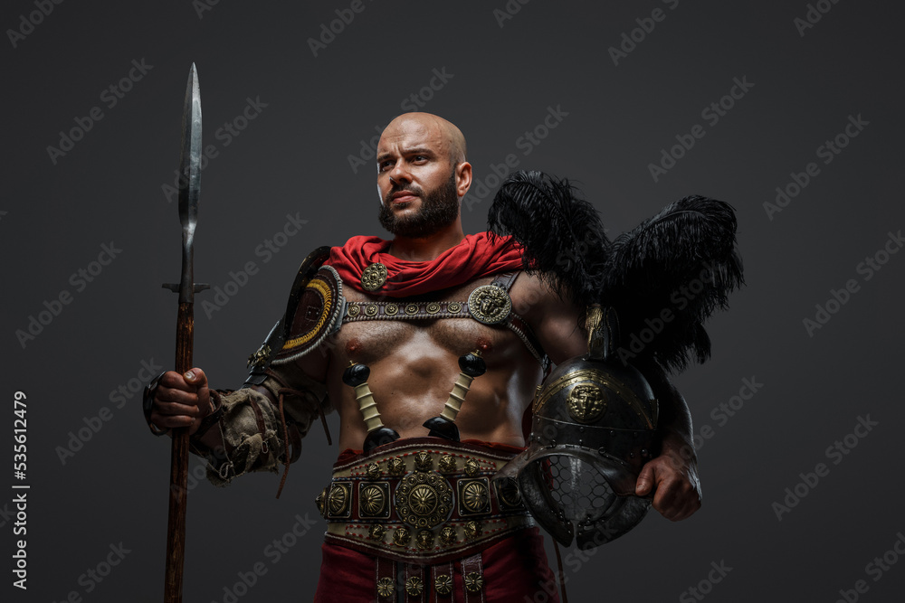Obraz premium Shot of powerful antique gladiator dressed in armor holding plumed helmet and long spear.