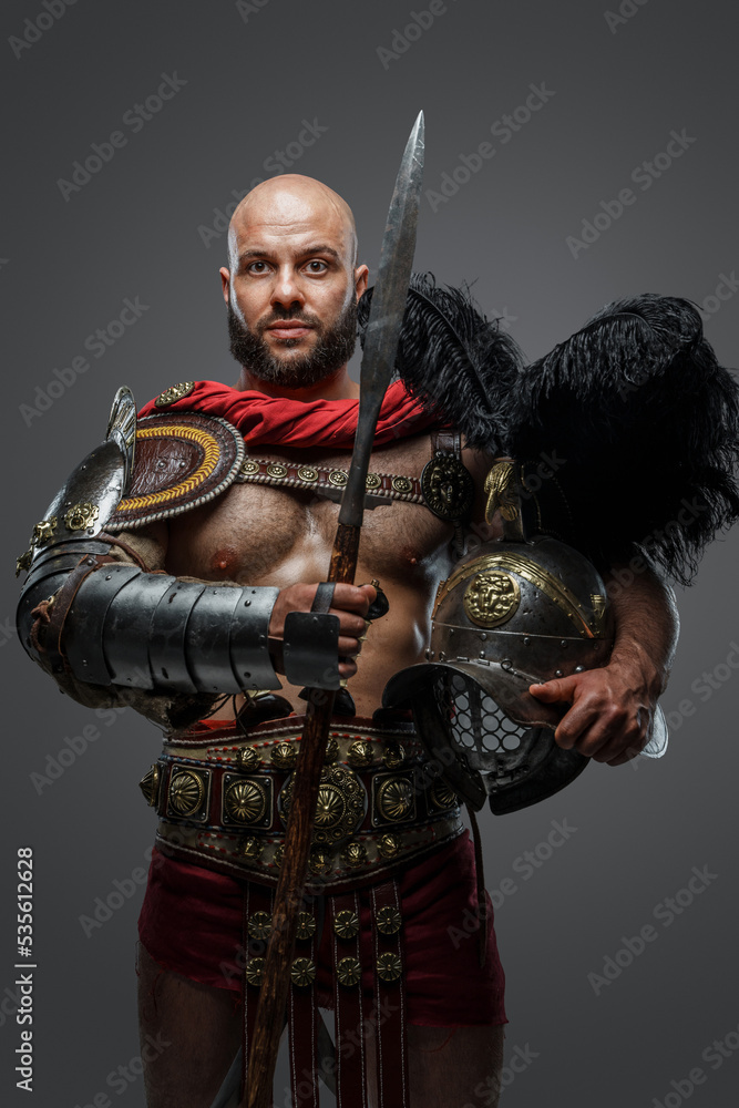 Fototapeta premium Shot of powerful antique gladiator dressed in armor holding plumed helmet and long spear.