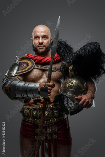 Shot of powerful antique gladiator dressed in armor holding plumed helmet and long spear.