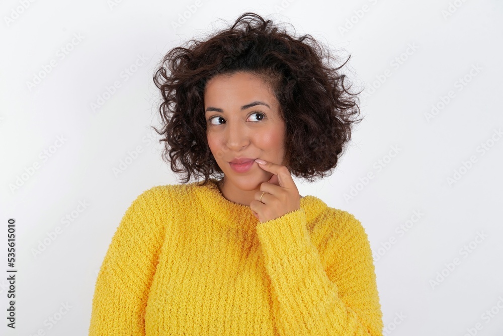 Lovely dreamy young beautiful woman with curly short hair wearing yellow sweater over white background keeps finger near lips looks aside copy space. Lovely dreamy young beautiful woman with curly short hair wearing yellow sweater over white background keeps finger near lips looks aside copy space.