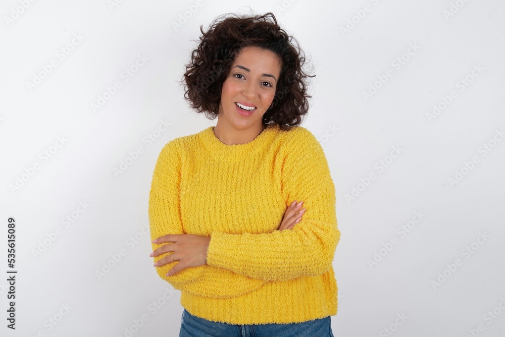 Portrait of charming young beautiful woman with curly short hair wearing yellow sweater over white background standing confidently smiling toothily with hands folded Portrait of charming young beautiful woman with curly short hair wearing yellow sweater over white background standing confidently smiling toothily with hands folded