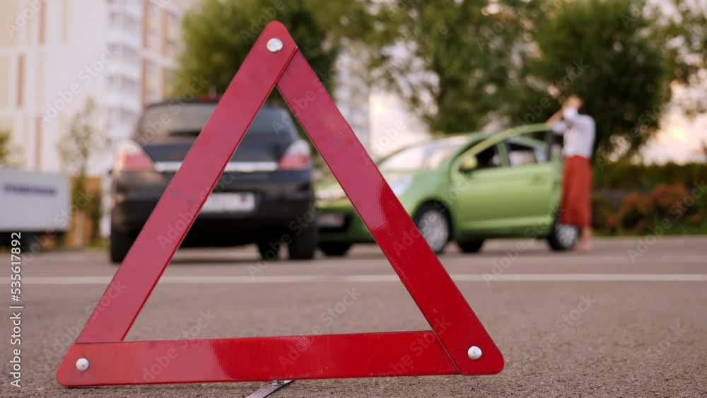 Close-up of a reflective red triangle on the road, with an accident in the background. Close-up of a reflective red triangle on the road, with an accident in the background.