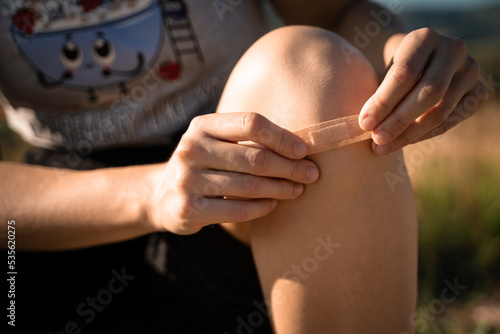 Close up of girl putting band-aid on knee after injury, fall, illness, protect wound.  Concept of health and healthcare, physical recovery, outdoor sports.