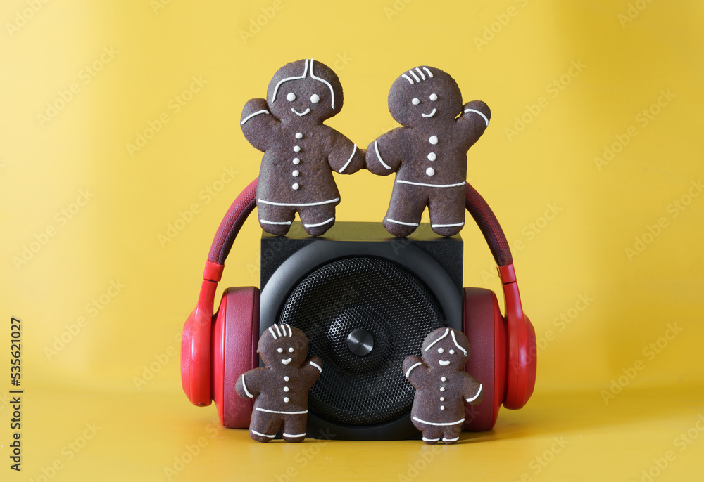 Black audio speaker with a metal grill, headphones and funny gingerbread men on a yellow background. Concept of a holiday party, pop music, family and children's audio broadcasts. Black audio speaker with a metal grill, headphones and funny gingerbread men on a yellow background. Concept of a holiday party, pop music, family and children's audio broadcasts.