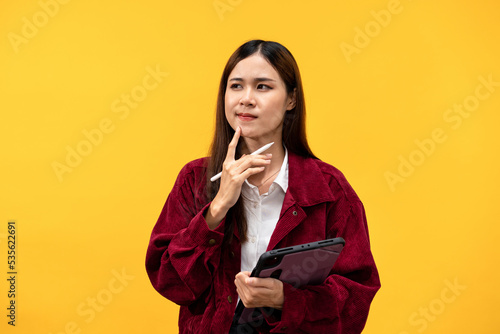 Young asian woman wearing red jacket and holding tablet with the pen to working while touching chin