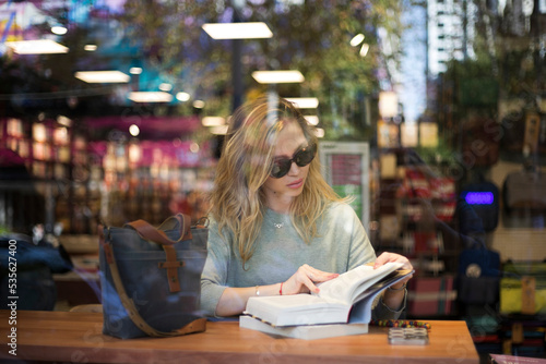 Photography View through the book shop window of young blonde woman reading book, wearing sunglasses