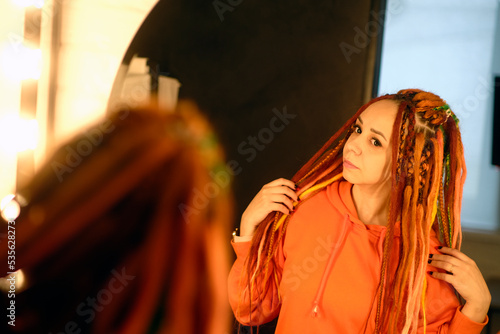 A beautiful woman in casual clothes and with dreadlocks looks in the mirror, standing in a modern apartment