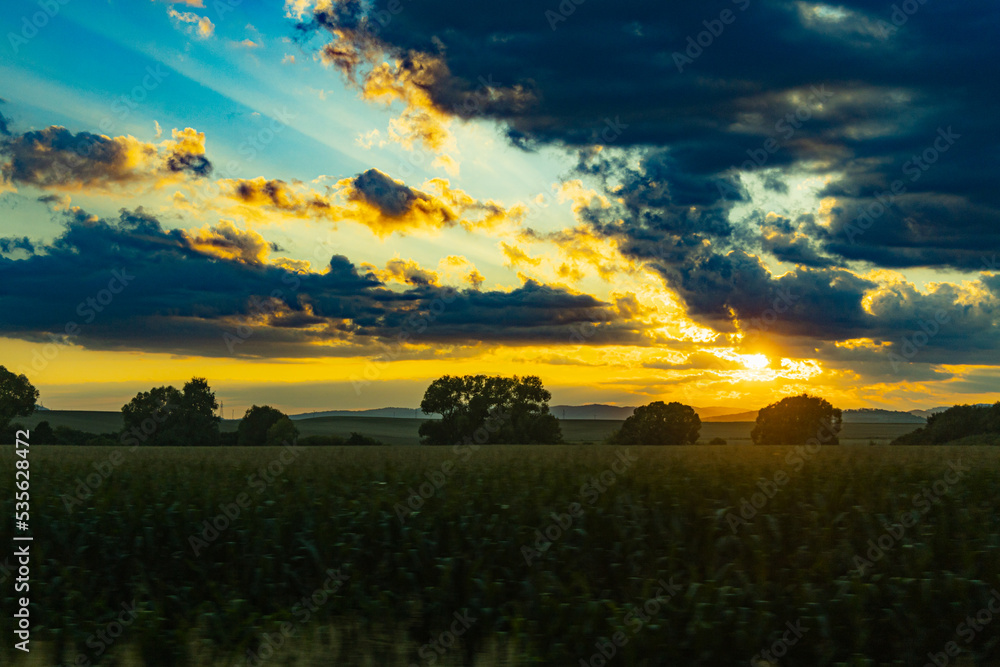 Naklejka premium Beautiful shining sun behind big clouds at sunset over big fields