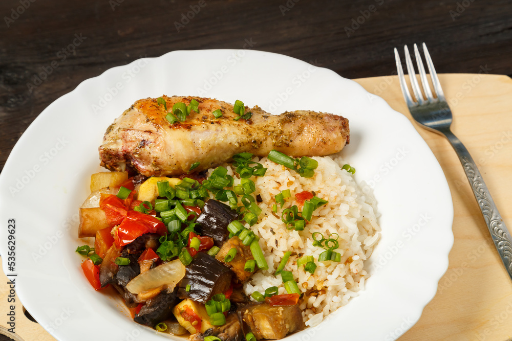 Rice with vegetables in sauce and chicken leg in a white plate on a handmade wooden board next to a fork on a wooden table. Rice with vegetables in sauce and chicken leg in a white plate on a handmade wooden board next to a fork on a wooden table.