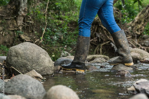 migrant woman crossing a stream wearing blue jeans and black marsh boots. hiking through the dense colombian forest. wet boots. hiking concept.