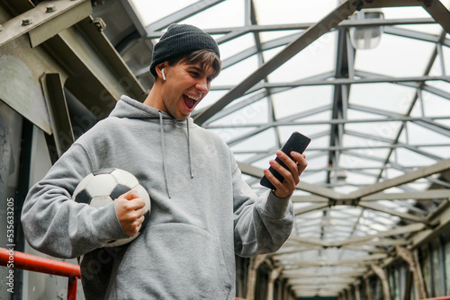 Foto Young hipster man in hoodie and hat holding smartphone and celebrate win a bet on the urban background