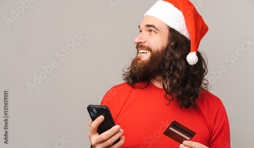 Cropped photo of cheerful bearded man wearing santa red hat holding smarphone and credit card and looking away