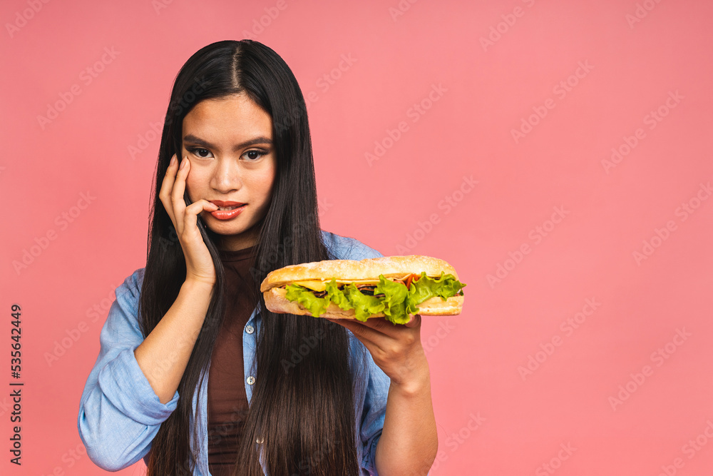 Young beautiful asian japanese chinese woman eating sandwich or big burger with satisfaction. Girl enjoys tasty hamburger takeaway, diet concept, standing isolated over pink background. Young beautiful asian japanese chinese woman eating sandwich or big burger with satisfaction. Girl enjoys tasty hamburger takeaway, diet concept, standing isolated over pink background.