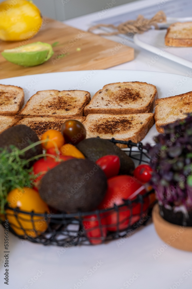 black metal basket with colorful vegetables, red and yellow tomatoes, avocado and green thyme and appetizing toasts on white kitchen table black metal basket with colorful vegetables, red and yellow tomatoes, avocado and green thyme and appetizing toasts on white kitchen table