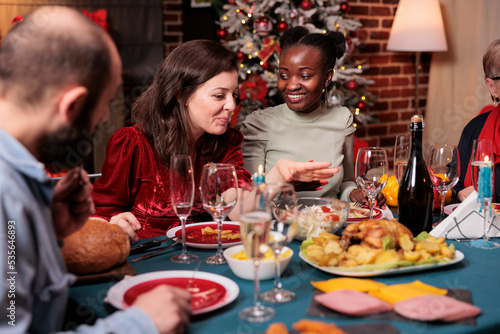 Diverse friends chatting at christmas home party, women talking at festive dinner table, people eating traditional xmas food. Winter holiday celebration in beautiful luxury decorated place