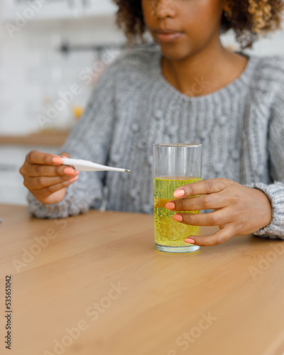 African American woman with a cold measures her body temperature and drinks soluble pills or vitamins, A tired sick woman at home takes pills for sore throat and viruses. treatment of influenza and