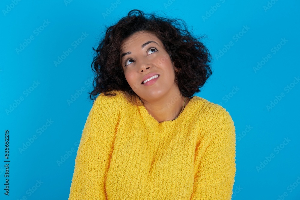 Portrait of mysterious young beautiful woman with curly short hair wearing yellow sweater over blue wall looking up with enigmatic smile. Advertisement concept. Portrait of mysterious young beautiful woman with curly short hair wearing yellow sweater over blue wall looking up with enigmatic smile. Advertisement concept.