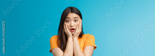 Close-up concerned shocked young timid asian girl gasping, dropping jaw feel sorry, pitty for friend open mouth stunned stare camera, grab face shook, speechless, frustrated, stand blue background photo