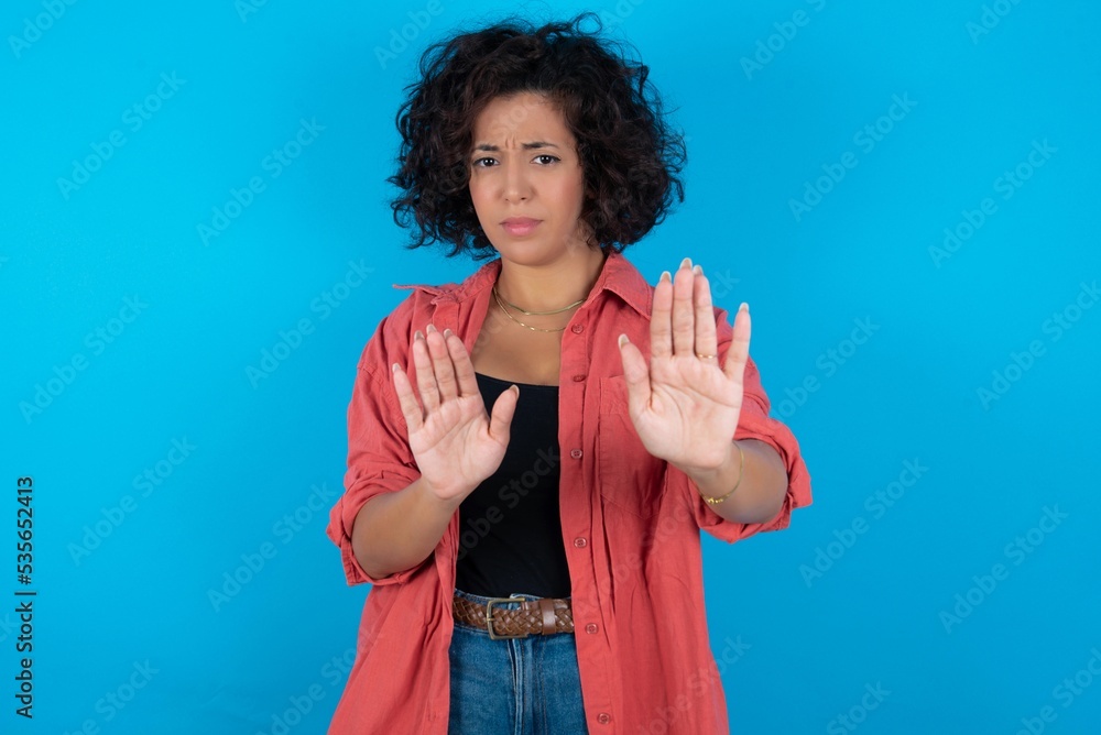 Afraid young beautiful woman with curly short hair wearing red overshirt over blue wall, makes terrified expression and stop gesture with both hands saying: Stay there. Panic concept. Afraid young beautiful woman with curly short hair wearing red overshirt over blue wall, makes terrified expression and stop gesture with both hands saying: Stay there. Panic concept.