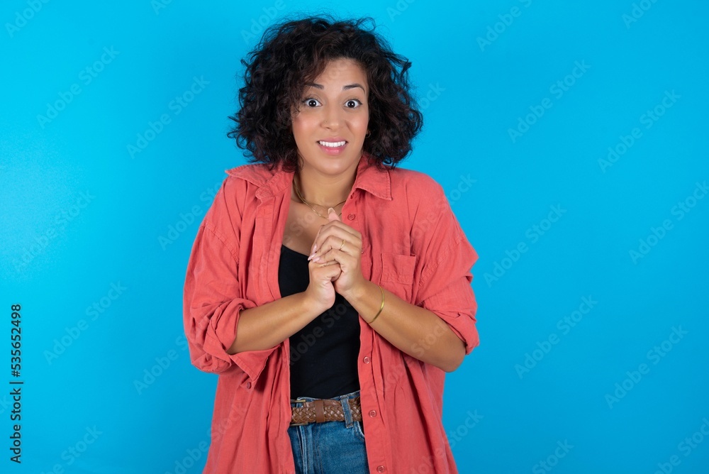 Happy young beautiful woman with curly short hair wearing red overshirt stands against orange studio wall keeps hands on heart, swears be loyal, expresses gratitude. Honesty concept. Happy young beautiful woman with curly short hair wearing red overshirt stands against orange studio wall keeps hands on heart, swears be loyal, expresses gratitude. Honesty concept.