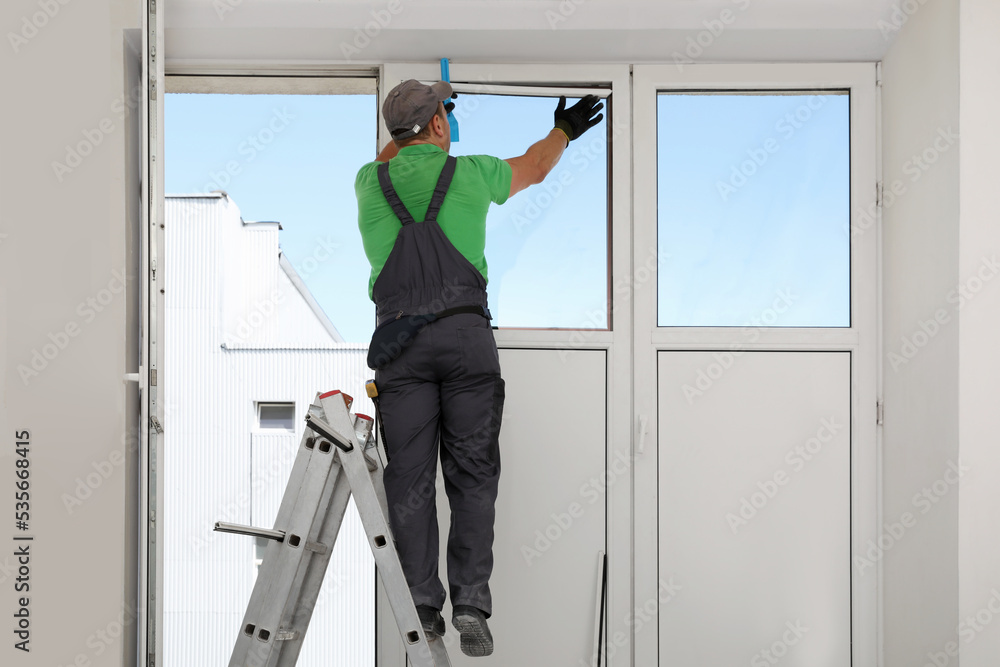 Worker on folding ladder installing window indoors, back view