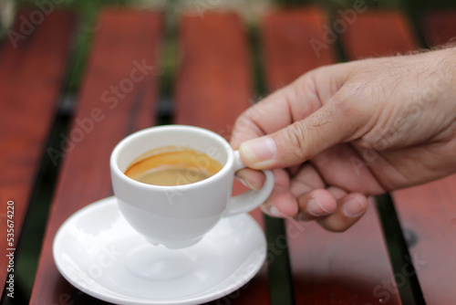 Hand holding espresso coffee in white ceramic cup on wooden table background. Espresso can be made with a wide variety of coffee beans and roast degrees.