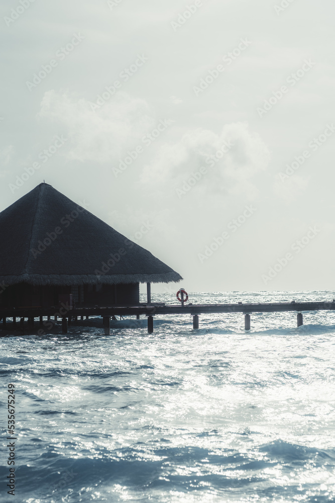 A vertical shot of a seascape with a canopy bungalow of a luxurious Maldives resort with a triangle roof and a wooden pier with a lifebuoy on it on a warm bright day with highlights on the waves