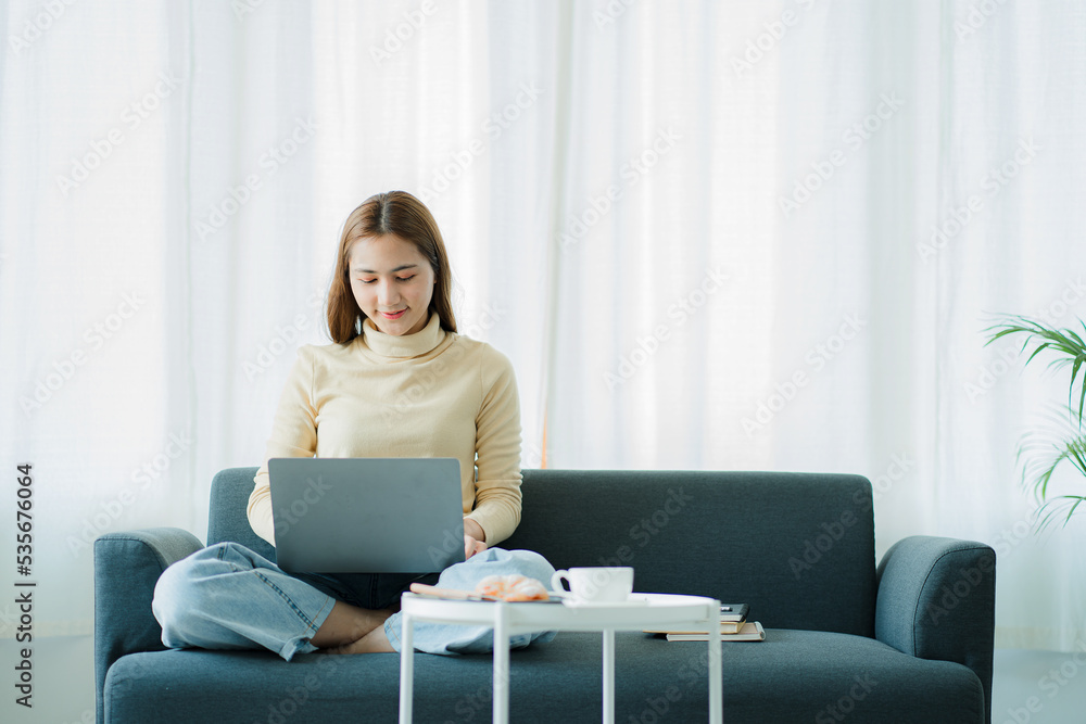 Asian woman sitting on the sofa with a laptopAttractive young asian woman using laptop computer while sitting on a couch at home Asian woman sitting on the sofa with a laptopAttractive young asian woman using laptop computer while sitting on a couch at home