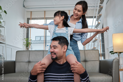 Asian Thai family together, dad plays and teases with daughter and mum by carrying and holding girl on shoulders in home living room, happy leisure times, lovely weekend, wellbeing domestic lifestyle.
