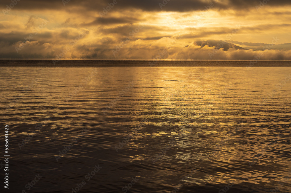 雲と遠くの靄がさざ波の水面と共に黄金色に染まる湖の風景。北海道の屈斜路湖。