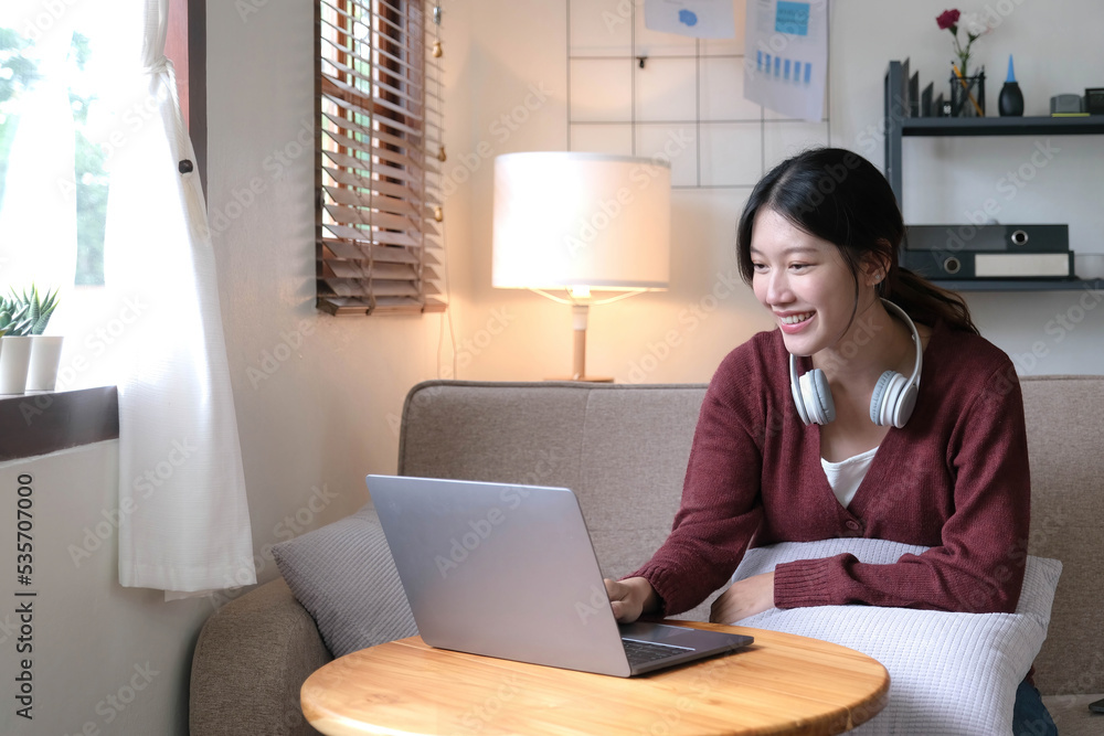 Beautiful Asian woman using laptop while headphones sitting on sofa at home Beautiful Asian woman using laptop while headphones sitting on sofa at home