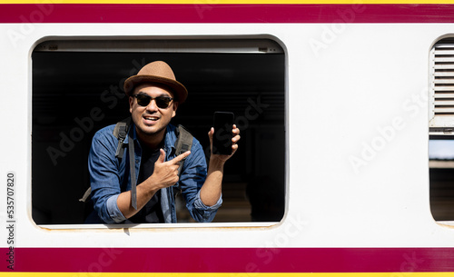 Young asian man travel by train. Sticking his head outta the train window showing smartphone. Backpacker arrival and departure at platform railway. Freedom  trip on vacation time holiday weekend. photo