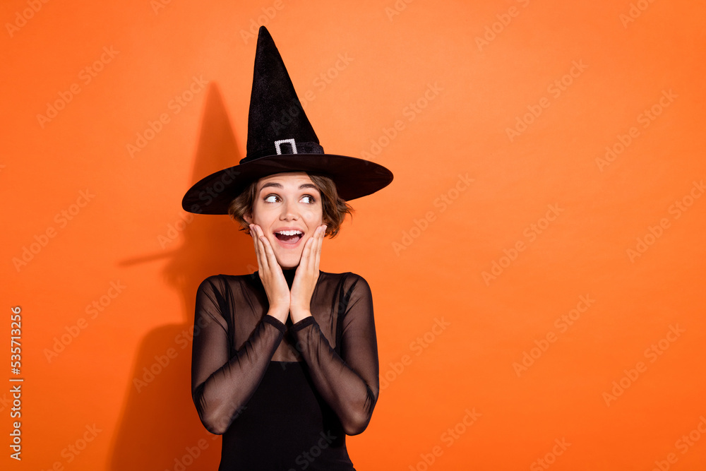 Photo of impressed funny lady wear black witch dress cap arms hands cheekbones cheeks looking empty space isolated orange color background Photo of impressed funny lady wear black witch dress cap arms hands cheekbones cheeks looking empty space isolated orange color background