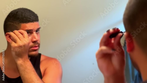 Gay mixed race man looking into mirror adding eyebrow makeup in bathroom, close up LGBTQ photo