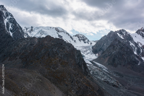 Atmospheric landscape with large snow mountain range with glacier and icefall in dramatic cloudy sky. Awesome high snowy mountains under rainy clouds. White snow on black rocks in overcast weather.