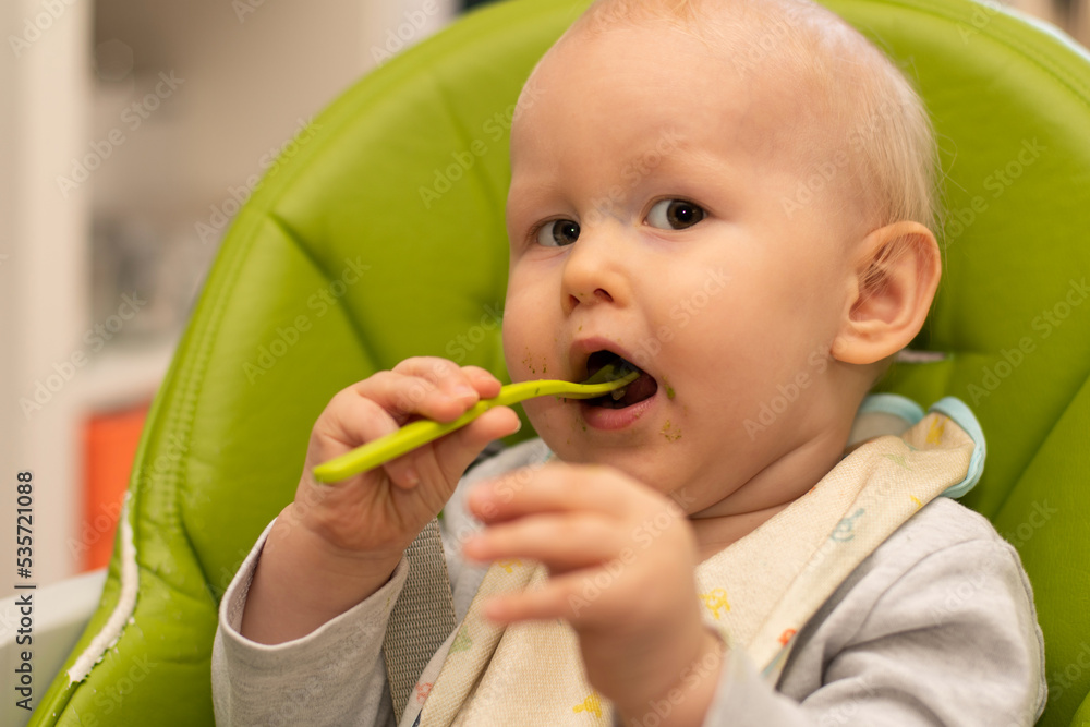 Little baby playing and feeding himself with spoon sitting in highchair. Baby first solids. Weaning. Little baby playing and feeding himself with spoon sitting in highchair. Baby first solids. Weaning.