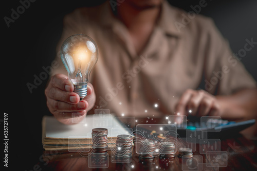 Businessman holding light bulb and using calculator to calculate, Stacking coins, graph, notebook, pen on table. plan revenue and expenses, target success growth, finance profit planning concept.