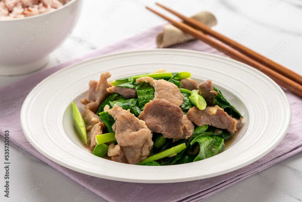 Stir fried pork slices with Chinese kale, a Thai dish presenting that Thai food also does reflect the Chinese cuisine, served on a white serving plate together with a cup of cooked brown jasmine rice. Stir fried pork slices with Chinese kale, a Thai dish presenting that Thai food also does reflect the Chinese cuisine, served on a white serving plate together with a cup of cooked brown jasmine rice.