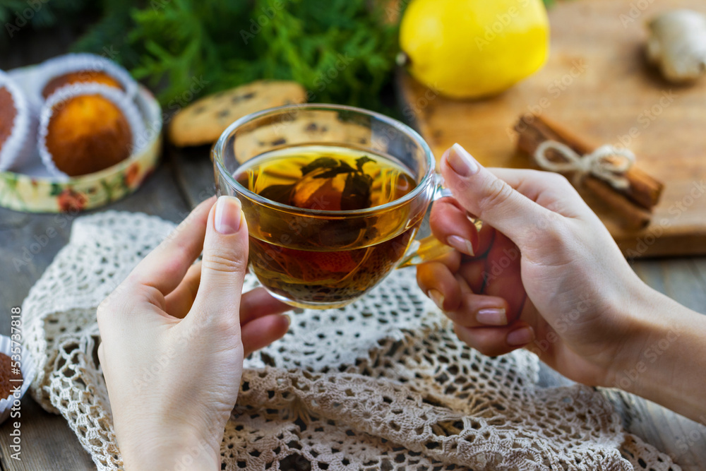 Top view of glass cup of tea with mint in female hand. Homemade chocolate chip cookies, lemon, cinnamon sticks, honey and thuja branches on wooden background Top view of glass cup of tea with mint in female hand. Homemade chocolate chip cookies, lemon, cinnamon sticks, honey and thuja branches on wooden background