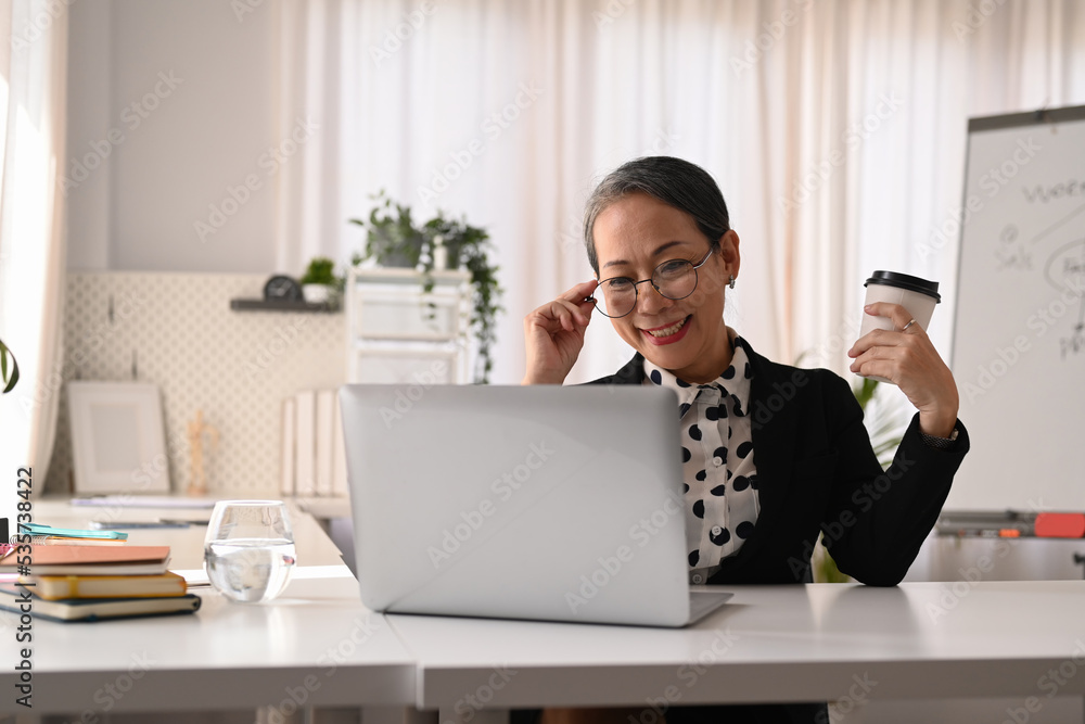 Smiling senior businesswoman wearing elegant formal suit using laptop computer in modern personal office Smiling senior businesswoman wearing elegant formal suit using laptop computer in modern personal office