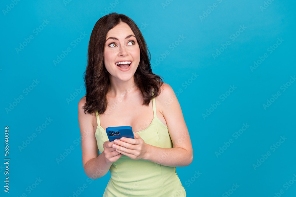 Photo of shiny excited girl dressed green clothes writing modern device looking empty space isolated blue color background Photo of shiny excited girl dressed green clothes writing modern device looking empty space isolated blue color background